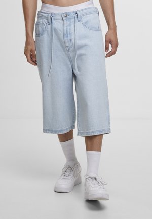 Mężczyzna noszący jasnoniebieskie szorty dżinsowe do kolan, białe skarpety i białe trampki, stojący na szarej podłodze na białym tle.