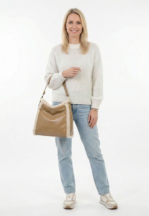 Blonde vrouw draagt een witte trui en lichte spijkerbroek, houdt een beige handtas met schapenvachtaccent vast, staand voor een witte achtergrond.