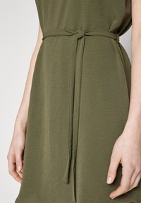 Olivově zelené šaty z texturované látky s opaskem zavázaným v pase, s jednoduchým a volným střihem. Viditelná ruka odpočívající vedle opasku.