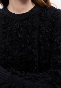 Gros plan sur un pull noir en maille avec des motifs en franges texturées, un col côtelé et des manches longues, porté par une personne à la peau claire et aux cheveux bruns.