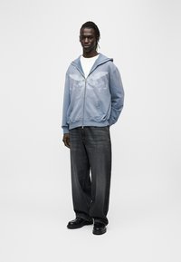 Μπλε φούτερ με φερμουάρ και γραφικό σχέδιο, συνδυασμένο με σκούρο τζιν και μαύρα παπούτσια. Το φούτερ διαθέτει κουκούλα με κορδόνι και μπροστινές τσέπες.