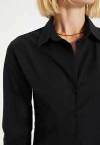 Donna con capelli biondi corti che indossa una camicia nera aderente con bottoni e una sottile collana d'oro su uno sfondo neutro.