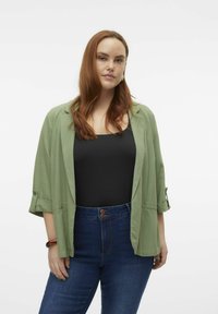 Veste légère verte avec manches retroussées, associée à un haut noir et un jean bleu taille haute. Texture douce et coupe décontractée.