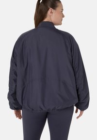 Frau mit braunen Haaren im Pferdeschwanz, die eine dunkelblaue, übergroße Jacke und passende Leggings trägt, steht mit dem Rücken zu einem schlichten Hintergrund.