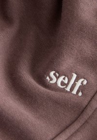 Lähedalt pruuni riide pildistus valge tikandiga tekstiga ""self."" tekstuurse, kergelt kortsus pinnaga.
