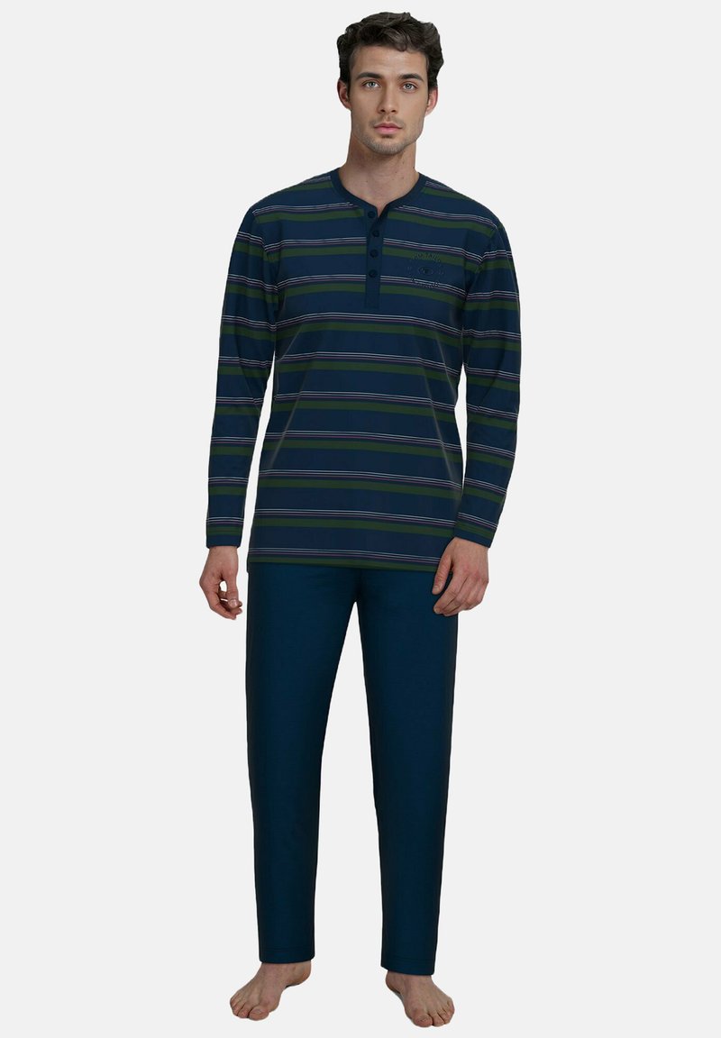 Ein Mann steht barfuß, trägt ein marineblaues, langärmliges, gestreiftes Henley-Shirt und einfarbige marineblaue Hosen vor einem schlichten grauen Hintergrund.