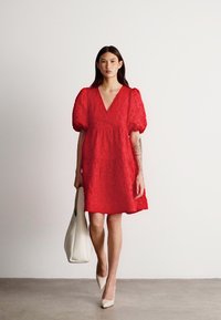 Raudona nėriniuota suknelė su pūkuotomis rankovėmis, V formos iškirpte, sluoksniuota sijonė; derinama su balta rankine ir smėlio spalvos aukštakulniais ant neutralios fono.