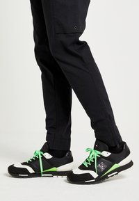 Černobílé atletické boty s zelenými akcenty a tkaničkami. Mají texturovaný svršek a kontrastní podrážku. Nosí se s tmavými kalhotami.