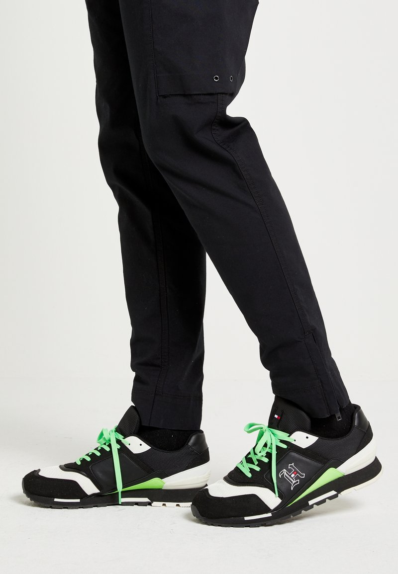 Černobílé atletické boty s zelenými akcenty a tkaničkami. Mají texturovaný svršek a kontrastní podrážku. Nosí se s tmavými kalhotami.