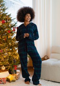 Groen- en marineblauw geruit pyjamaset met lange mouwen, een knoopsluiting aan de voorkant en rode accenten, gecombineerd met blote voeten in een gezellige kamersetting.