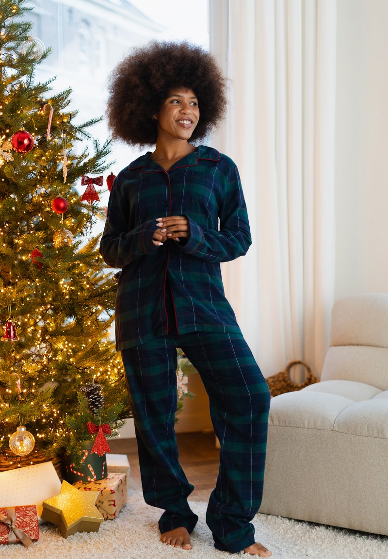 Groen- en marineblauw geruit pyjamaset met lange mouwen, een knoopsluiting aan de voorkant en rode accenten, gecombineerd met blote voeten in een gezellige kamersetting.