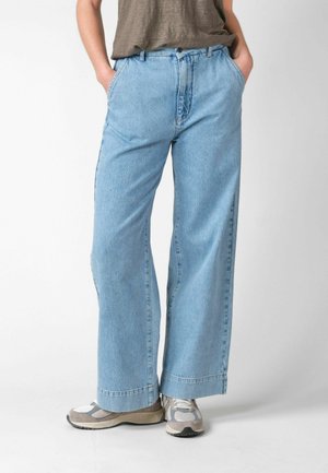 Person iført lyseblå, vidde bukser med hænderne i lommerne og hvide og grå sneakers, stående mod en ensfarvet baggrund.