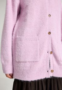 Lyserød cardigan lavet af blødt, tekstureret materiale. Har runde træknapper og to forlommer. Ribstrikkede manchetter og kant.