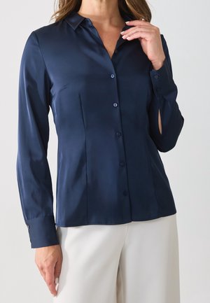 Femme portant une chemise boutonnée ajustée bleu marine à manches longues, associée à un pantalon couleur crème, main près du col, fond neutre.