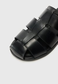 Black leather sandal with an open toe design, featuring woven straps and a subtle sheen. Soft texture with a durable sole.
