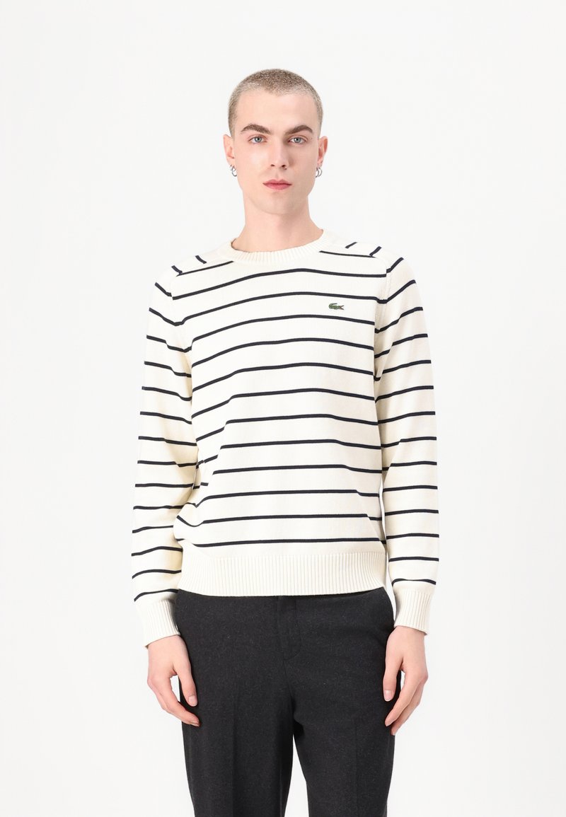 Sweater creme com riscas azuis, gola redonda, punhos e bainha canelados, apresentando um pequeno logótipo verde no peito. Usado com calças escuras.