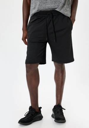 Pantalones cortos deportivos negros hechos de un tejido suave, con una cintura elástica y cordones, además de bolsillos laterales. Combinados con zapatillas negras.