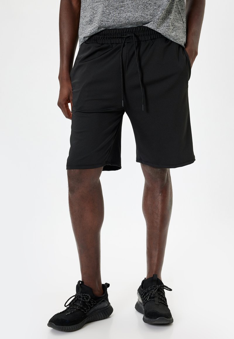 Pantalones cortos deportivos negros hechos de un tejido suave, con una cintura elástica y cordones, además de bolsillos laterales. Combinados con zapatillas negras.