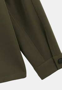 Poignet de chemise à manches longues vert foncé avec bouton et couture détaillée sur fond neutre.