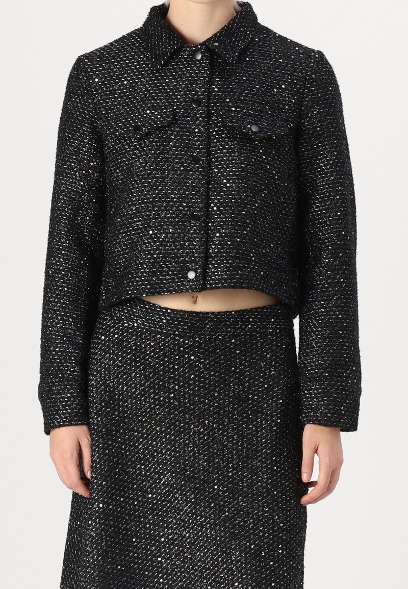 Femme portant une veste courte texturée noire avec des boutons et une jupe assortie, laissant apercevoir une petite partie du ventre.