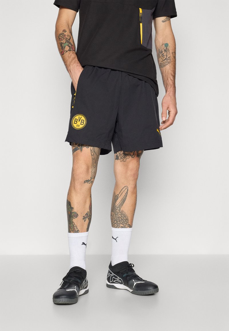 Sorte sportshorts med et gult Borussia Dortmund-logo, letvægtsmateriale og løs pasform. Bæres med hvide sokker og sorte sportssko.