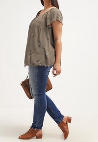 Topsi tekstuurit, oliivinvihreä, lyhythihainen ja kukka-applikaatioilla varustettu, yhdistettynä keskipitkiin sinisiin slim-fit-farkkuihin ja ruskeisiin avokkaisiin.