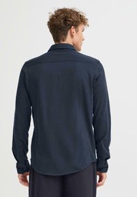 Mørkeblå langermet skjorte med knappede mansjetter, lett buet kant og en jevn tekstur. Har standard krage og bakstykke.