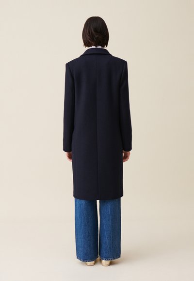 Cappotto di lana blu navy con un design strutturato, maniche lunghe e una fessura posteriore singola, abbinato a jeans di denim a gamba larga.