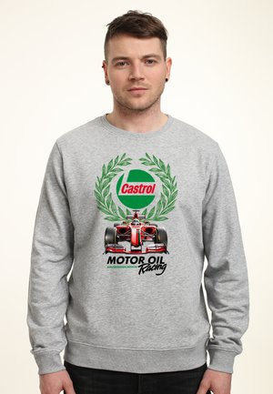 Młody mężczyzna w szarej bluzie z kapturem z logo Castrol, zielonym wieńcem laurowym, czerwonym samochodem Formuły 1 oraz napisem "Motor Oil Racing" na przodzie.