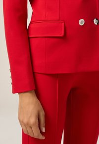 Czerwony dopasowany żakiet z klapami, z kieszenią i srebrnymi guzikami. Gładka, strukturalna tkanina o wyrazistym wykończeniu.