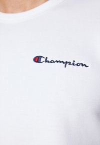 T-shirt en coton blanc avec un logo "Champion" brodé en marine et rouge sur la poitrine. Col rond à texture côtelée.