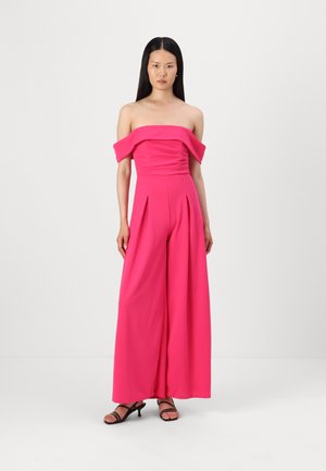 Jumpsuit rose vif décolleté avec des jambes larges plissées et un corsage ajusté. Tissu lisse et matériel minimal. Porté avec des sandales à talons noires.