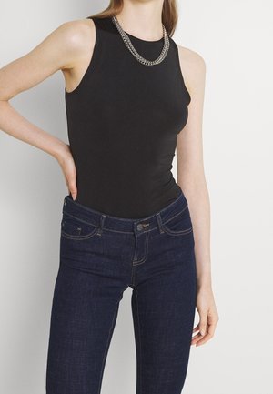 Kobieta ubrana w czarny, bezrękawnik, ciemnoniebieskie dżinsy oraz srebrny naszyjnik z łańcuszka, pozująca z jedną ręką na biodrze.