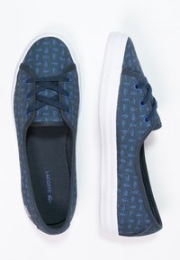 Mörkblå canvas skor med en texturerad ovandel med ett subtilt mönster, vit gummisula och mörka snören. Lacoste-logotyp på sidan.