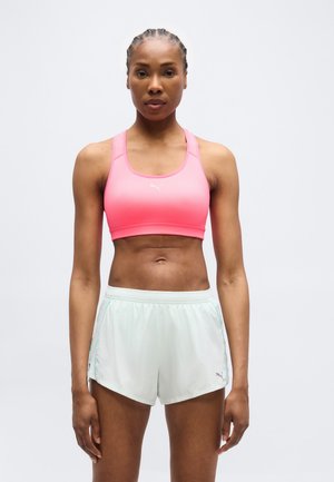 Femeie atletica cu părul împletit, purtând un sutien sport roz și pantaloni scurți de alergare Puma albi, stând în picioare pe un fundal simplu.