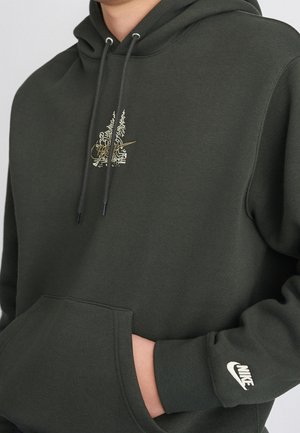 Person trägt dunkelgrünen Nike-Hoodie mit kleinem Grafikmotiv auf der Brust und weißem Nike-Logo am linken Ärmelbündchen, Hand in der vorderen Tasche.