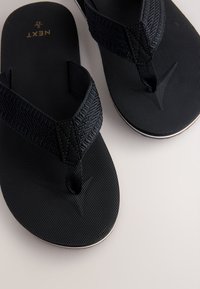 Tongs noires avec des lanières texturées et une semelle lisse, présentant un léger accent blanc autour de la base. Logo de la marque visible.