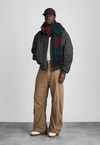 Schwarze Bomberjacke über einem weißen Hemd, beige weite Hose, grün-rot gemusterter Schal und braune Mütze, ergänzt durch weiße Schuhe.