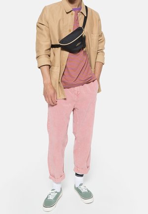 Person trägt beige Jacke, lila gestreiftes Hemd, pinke Hose, grüne Turnschuhe, weiße Socken und schwarze Umhängetasche vor weißem Hintergrund.