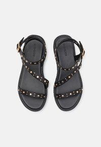Sorte flate sandaler med flere stropper, med gullstuds langs sidene, justerbar ankelrem og en flat, polstret såle.