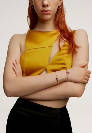Top de satén amarillo con un diseño recortado, combinado con una falda negra. Pulsera de cadena dorada con un charm púrpura en una muñeca.