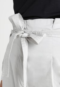 Witte stoffen riem die in een strik om de taille is geknoopt, met gestructureerde strepen en een gladde afwerking, bevestigd aan een lichte kledingstuk.