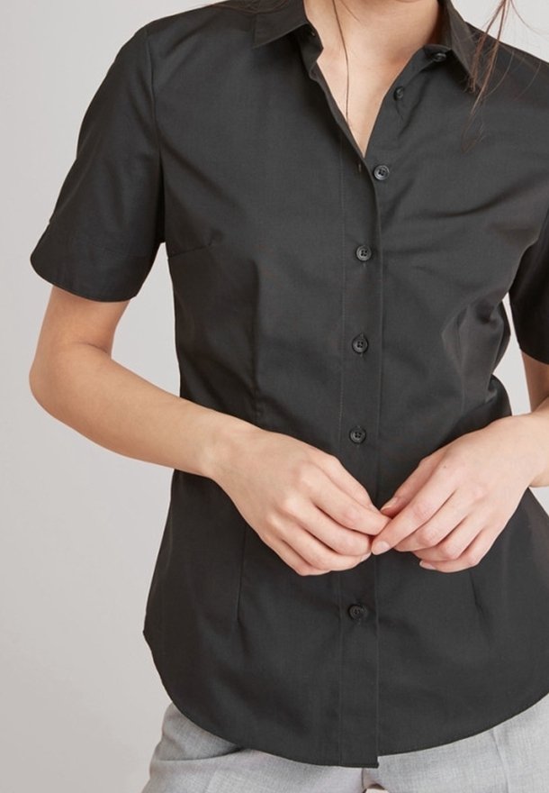 Femme portant une chemise noire ajustée à manches courtes boutonnée et un pantalon gris clair, les mains jointes à la taille.