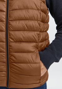 Braune isolierte Weste mit Steppmuster, ausgestattet mit einem schwarzen Reißverschluss und einem gerippten, marineblauen Ärmelsaum. Texturiertes, leichtes Material.