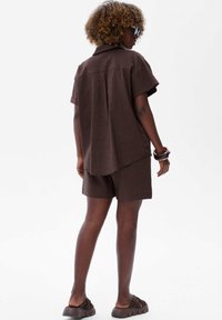 Kobieta z kręconymi włosami, ubrana w ciemnobrązową luźną koszulę z krótkim rękawem i pasujące szorty, masywne sandały i bransoletki, stojąca na tle białym.