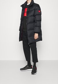 Abrigo acolchado negro con cuello alto y diseño acolchado; detalles y cremallera en rojo. Suéter negro con estampado rojo, pantalones negros y zapatillas.