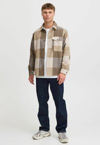 Giacca camicia a quadri marrone e beige con taschino, bottoni beige, indossata sopra una maglietta bianca, abbinata a jeans scuri e scarpe.
