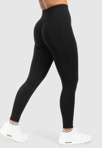 Persoon in zwarte high-waisted leggings en witte sneakers, geposeerd met één been naar achteren gestrekt tegen een eentonige lichte achtergrond.