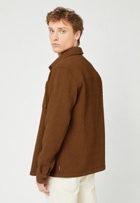 Junger Mann in einer strukturierten braunen Jacke und hellen Hosen, der über seine Schulter gegen einen schlichten hellen Hintergrund schaut.