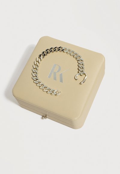 Bracelet en chaîne argentée avec un fermoir cassé reposant sur une boîte à bijoux beige avec les lettres "RK" argentées sur le dessus.